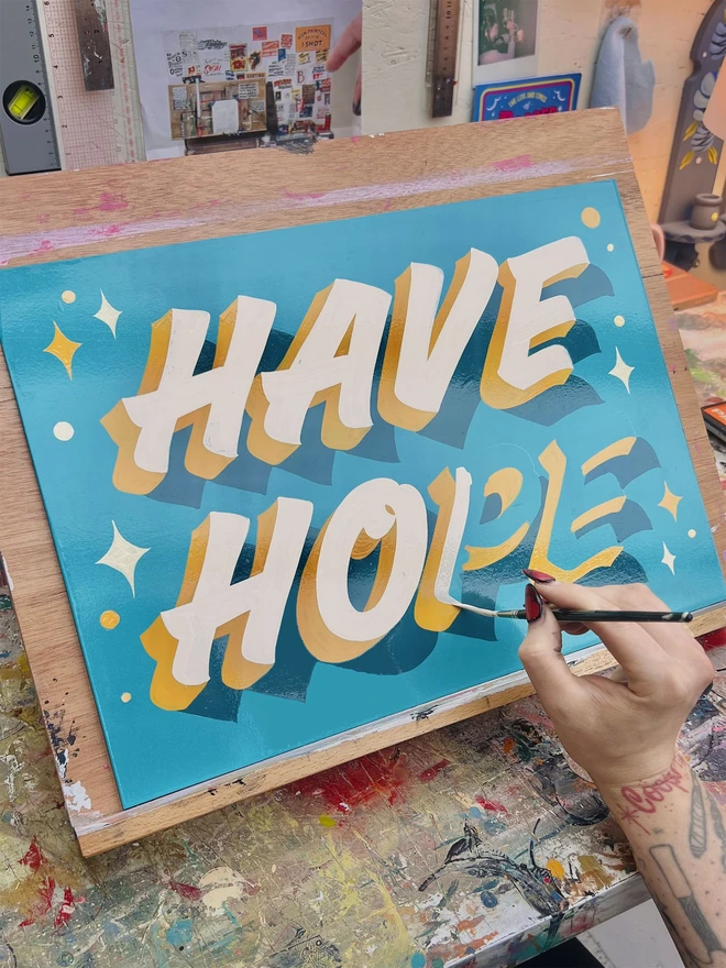 Woman's hand painting a sign saying 'Have Hope' in ivory and mustard colours on blue background with sparkly details.