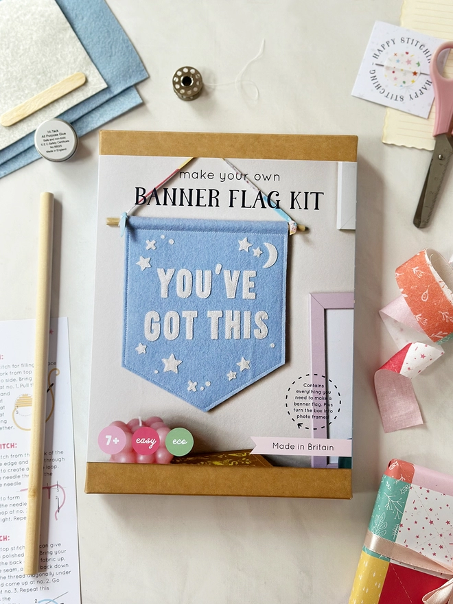 A craft kit box to make a felt positive message banner lays on a white desk with the components surrounding the box.