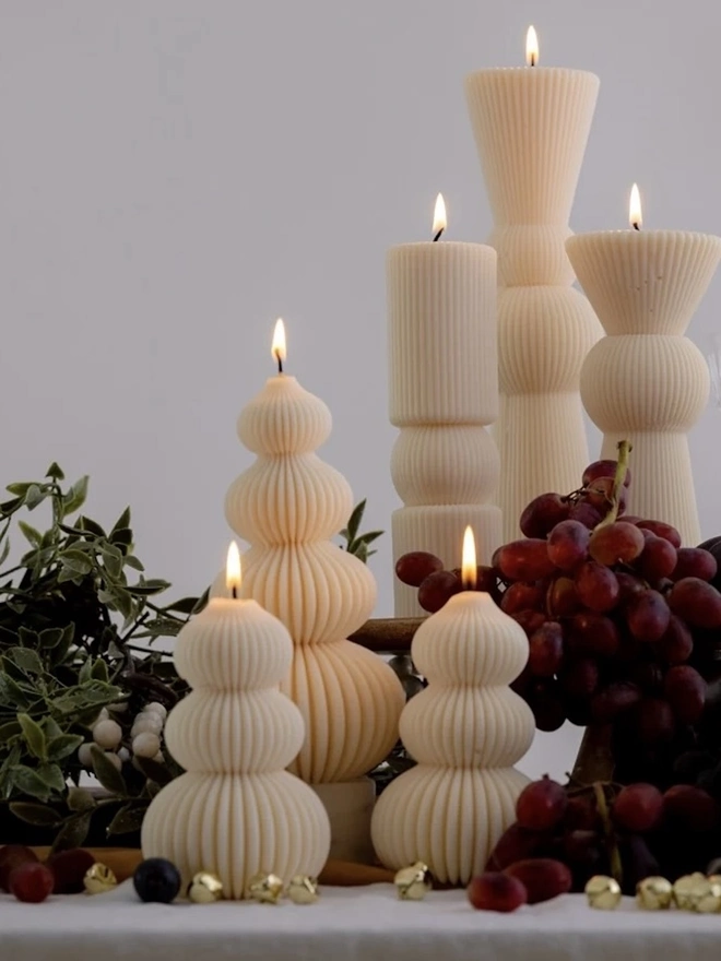 Signature Christmas Tree Sculpture Candle. Lit cream ribbed candles in stacked tree and totem shapes, styled on a festive table with red grapes, greenery and gold jingle bells. The sculptural candles make an elegant centrepiece for Christmas dinners and holiday gatherings.