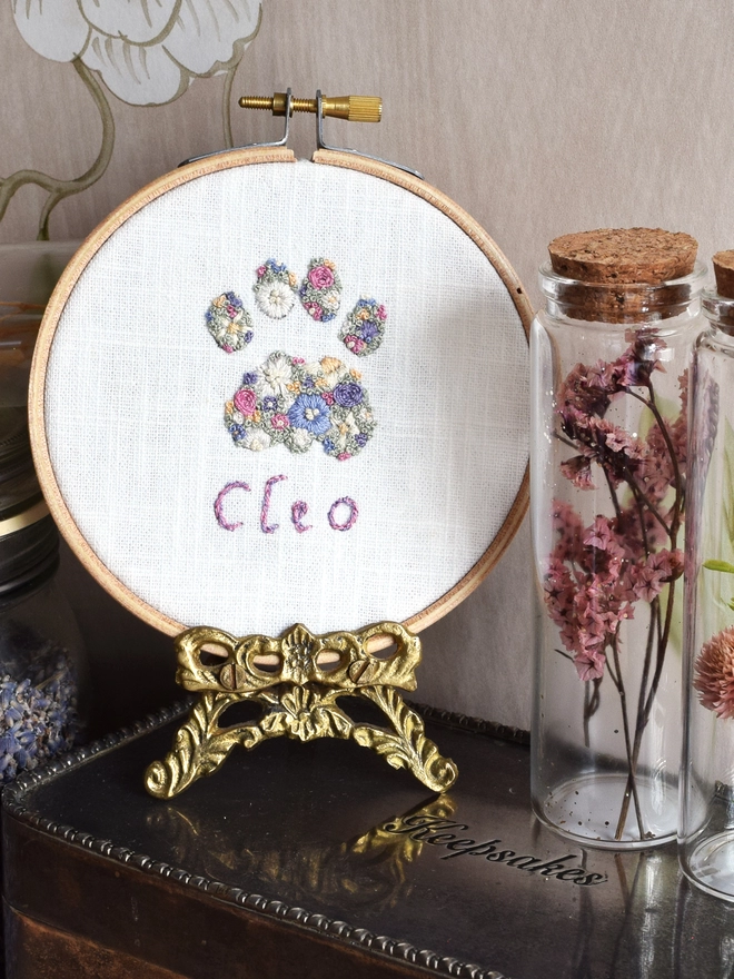 An embroidered Cats Paw, of Lavender Blues and Buttermilk yellow blossoms.  Displayed in a hoop frame on a Gold tone stand, on top of a tarnished silver keepsake tin with floral specimen glasses.