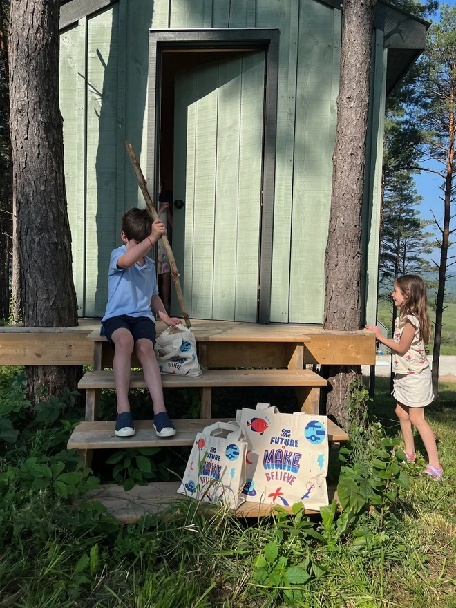A boy and a girl playing with canvas tote bags in the woods outside a tree house