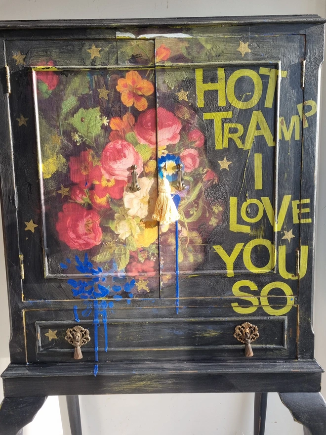 Elegant cocktail cabinet in black with colourful florals and text in chartreuse green that reads 'hot tramp, I love you so', a David Bowie lyric. Heavily distressed to age it. The interior is cobalt blue.