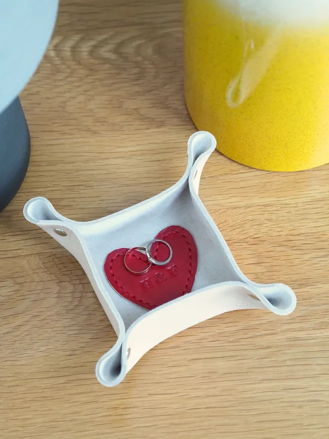A handmade mini leather square trinket tray with a red love heart shape stitched to the inside sitting on a side table. The tray has two wedding rings sitting inside for safe keeping.