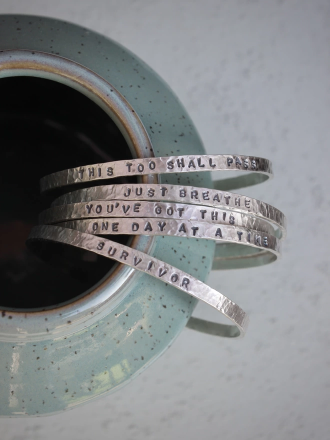 A selection of sterling silver cuff bangles with phrases stamped on them, arranged on the edge of a green ceramic vase on a grey background.