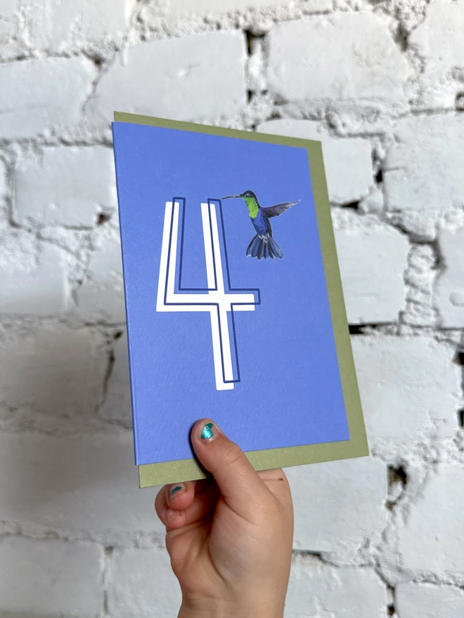 a deep purpley blue card with a big white number 4 and a hummingbird illustration and a green envelope