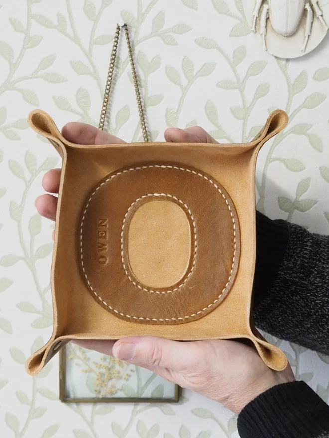 Handmade leather coin tray with a large letter decal stitched to the inside of the tray. A large letter O tan Brown coin tray held up by two hands to show the square shape and overall size of the valet tray.