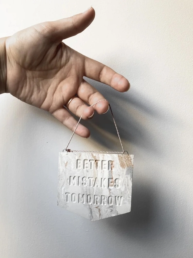 'Better Mistakes Tomorrow' Banner, a clay banner hanging against a plain backdrop, held by a person. 