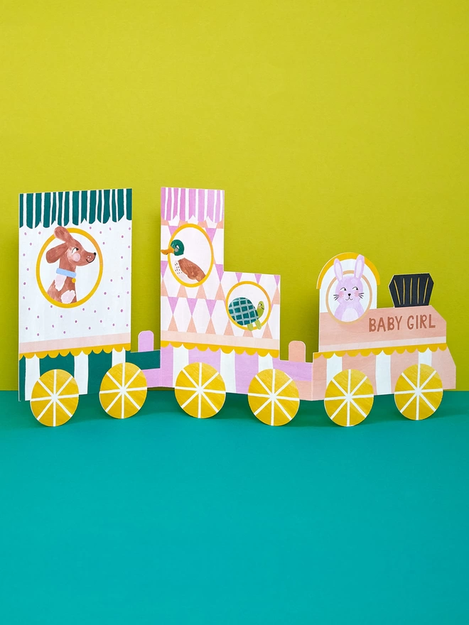 The concertina 'Baby Girl' fold-out card is a train with three carriages in patterned vibrant pink, peach, cream and green. Each carriage contains a character looking out the window, including a dog, duck, tortoise and pink rabbit