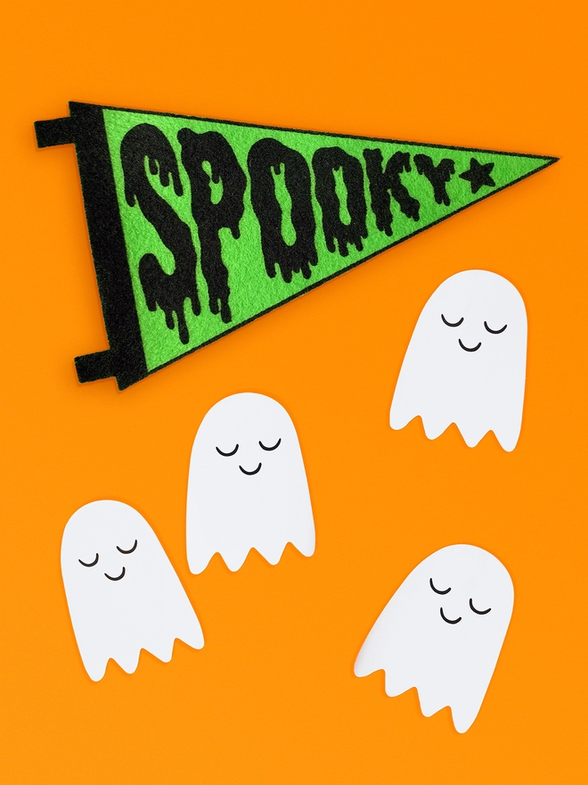Image shows a vibrant orange background with 4 small white paper ghosts dotted about, each ghost has a happy smiling face. Above the ghosts is a small felt pennant with the word ‘Spooky’ printed in black ink on bright green felt. The letters of the word are designed to look all gloopy and melty like the words is dripping. 