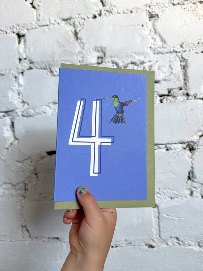 a deep purpley blue card with a big white number 4 and a hummingbird illustration and a green envelope