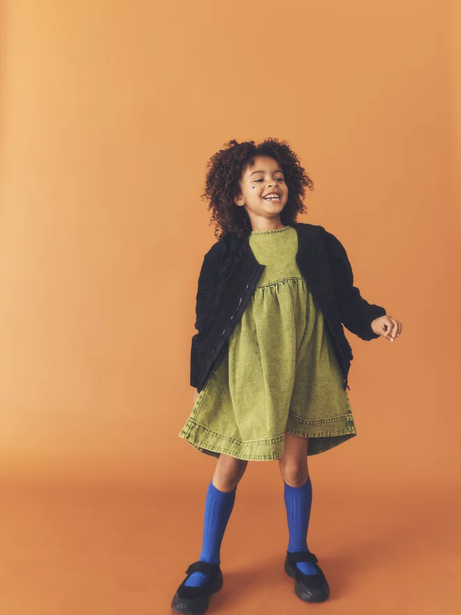 Child laughs facing camera against a burnt orange backdrop, wearing the Studio Koter Rumi dress in green acid-wash denim with cobalt knee socks.