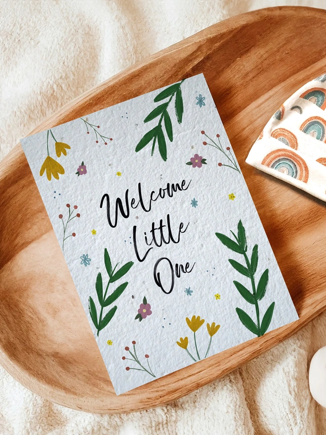 Floral Welcome Little One card on a wooden tray surrounded by blankets and a rainbow baby hat