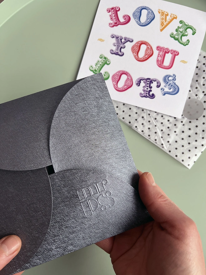Textured pouchette envelope containing a Mr.PS silk square, held in a pair of white hands