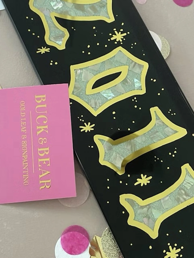 Personalised Year Date Sign, a black and gold personalised year sign sitting on a plain surface. It is joined by a small pink card.