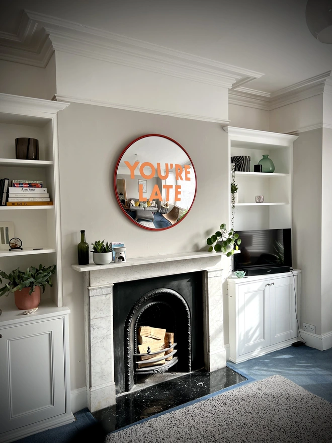 Round convex mirror with 'You're Late' handpainted in orange in a red round frame, above a cream fireplace surrounded by house plants