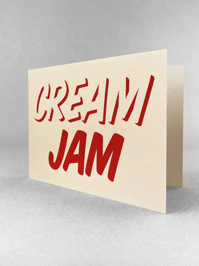 Jam on the bottom, Cream on top, printed in red ink on this cream card. Stood at an angle in a light grey studio with soft shadows.