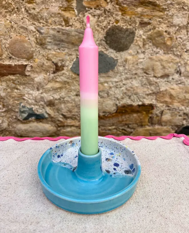 Shoreline Candle Stick Holder, a colourful candle stick holder sitting on a napkin, on a wooden surface against a brick backdrop. 