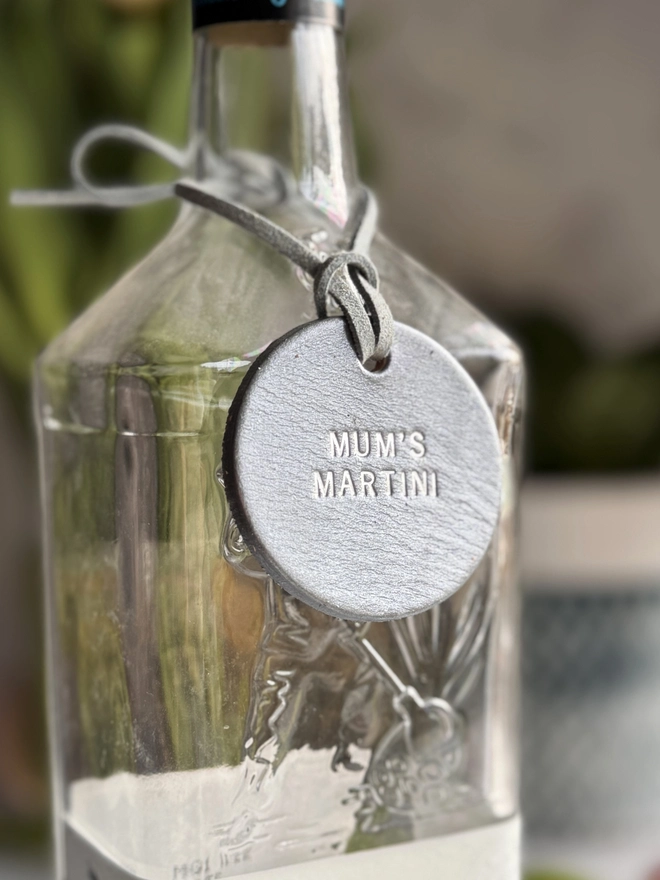 A glass bottle with a pewter leather tag hanging around the neck by a leather cord. The tag is hand-stamped with 'MUM'S MARTINI' in gold lettering, with pink tulips and a rustic background. A perfect personalised Mother’s Day gift or home bar accessory