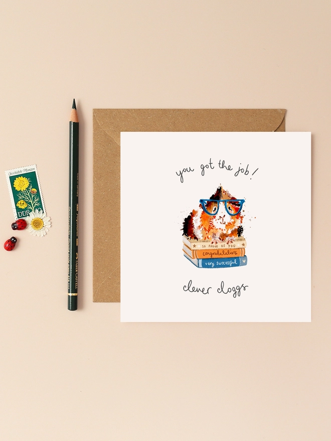 A whimsical illustration of a guinea pig wearing oversized glasses, perched atop a stack of books titled "Success Stories." The guinea pig gazes confidently ahead, embodying a sense of accomplishment. Above the scene, the playful, handwritten message "You got the job! Clever Clogs" adds a touch of humor and warmth. The design combines soft colors and charming details, making it a delightful way to congratulate someone on their new job.
