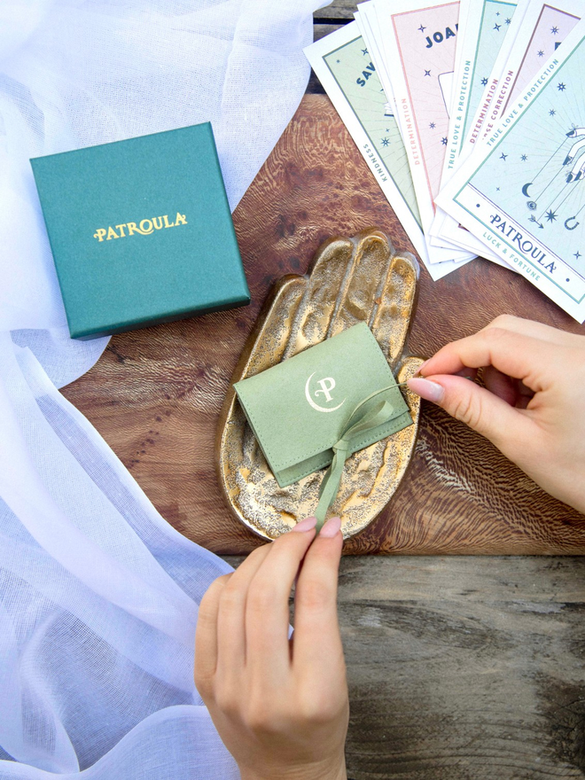 Hands tying a pale green jewellery pouch on a table with a dark green Patroula Jewellery box in the background