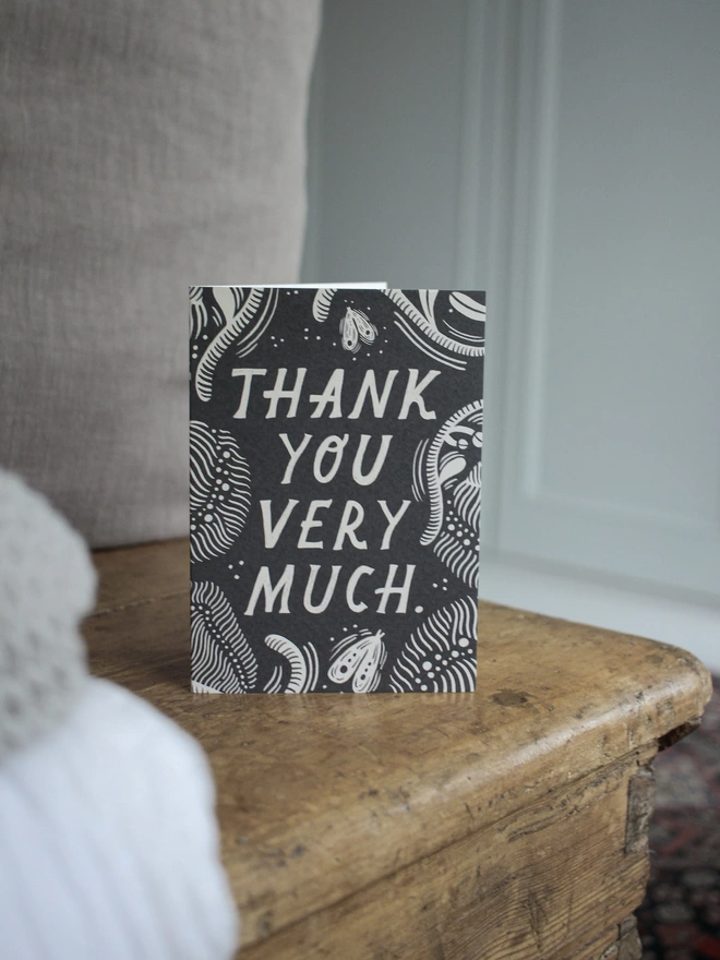 Black and white greeting card with illustration and the words thank you very much written on it stood up on a wooden surface with a grey cushion behind it 