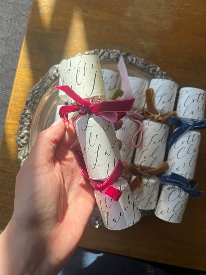 Selection of crackers personalised with calligraphy names tied with green, pink, mustard, blue, teal, rose, cerise velvet ribbon, laid on a silver tray on a wooden table