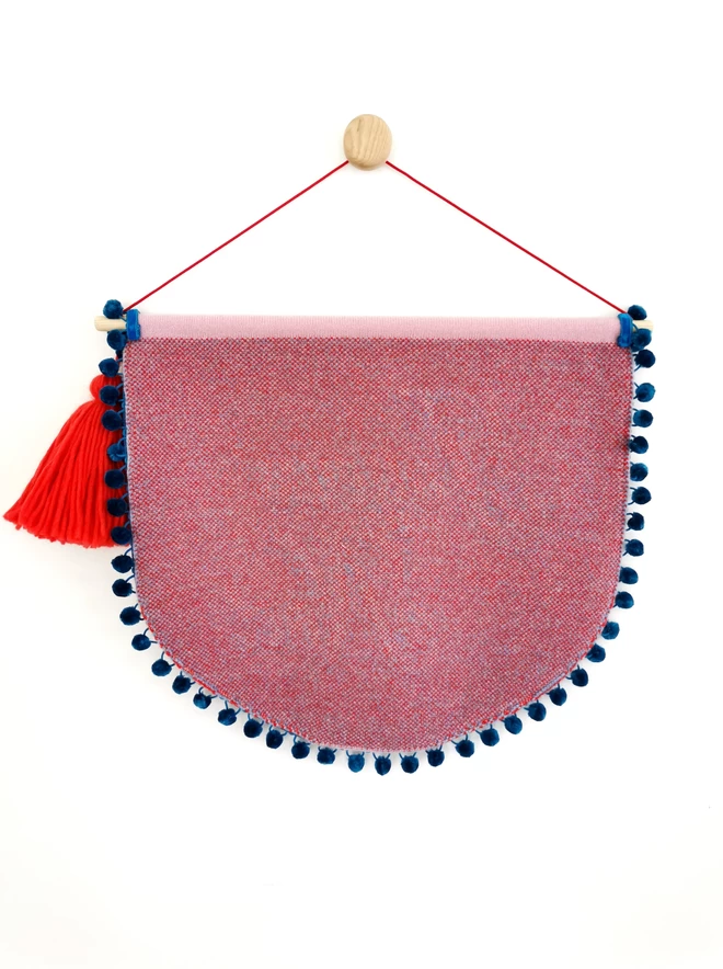 A product image showing the reverse of a scallop shaped wall hanging with teal pom pom trim edging. The back of the banner  is knitted into a red and blue birdseye check and an oversized red tassel can be seen hanging from one side of the wooden doweling. The whole banner is suspended from a wooden wall dot by a bright red nylon cord.