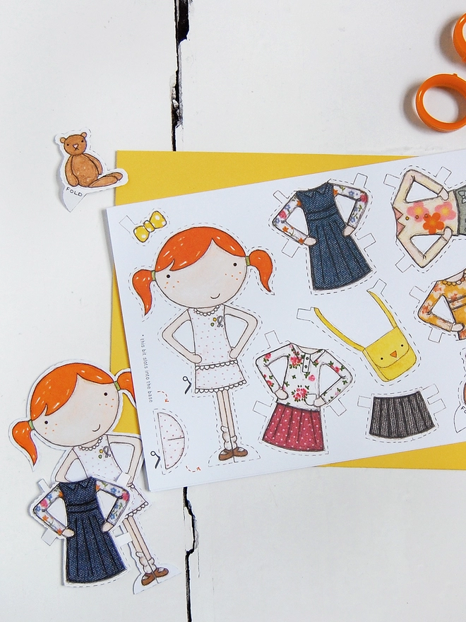 A paper doll greetings card with a paper doll and several outfits on the card lays on a yellow envelope on a white desk.