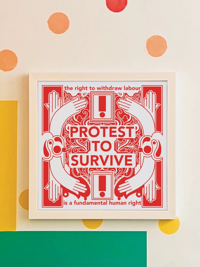 The bold red and white print with "Protest to survive" written at the centre, as well as “the right to withdraw labour” at the top, and “is a fundamental human right” at the bottom, is hanging in a white square frame on a white wall with yellow, orange, green and blue spots, and a green and yellow rectangle painted in the bottom left hand corner.