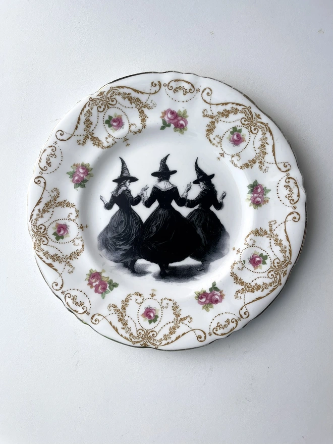 a coven of 3 witches dance hand in hand, the image is victorian in black and white and is printed directly onto an ornate vintage plate