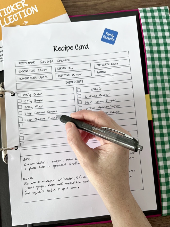 Close-up of hand writing on printed recipe card in an open green gingham recipe folder. Blue, square 'Family Favourite' sticker on page & yellow 'sticker collection' envelope peeking from behind page.