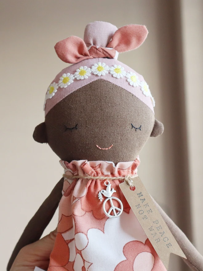 Handmade fabric doll called Peace Wishing Flower Child Doll, wearing peach and white retro floral dresses, daisy headbands, and bow topknots. It has serene embroidered face, peace sign charm around its neck, and tags reading “Make Peace Not War.” 