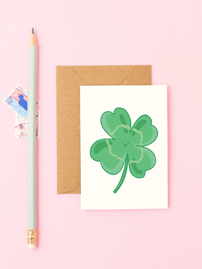 Mini greeting card featuring a hand-drawn four-leaf clover with a smiling face, styled with a kraft envelope, pastel green pencil, and vintage stamps on a pink background.