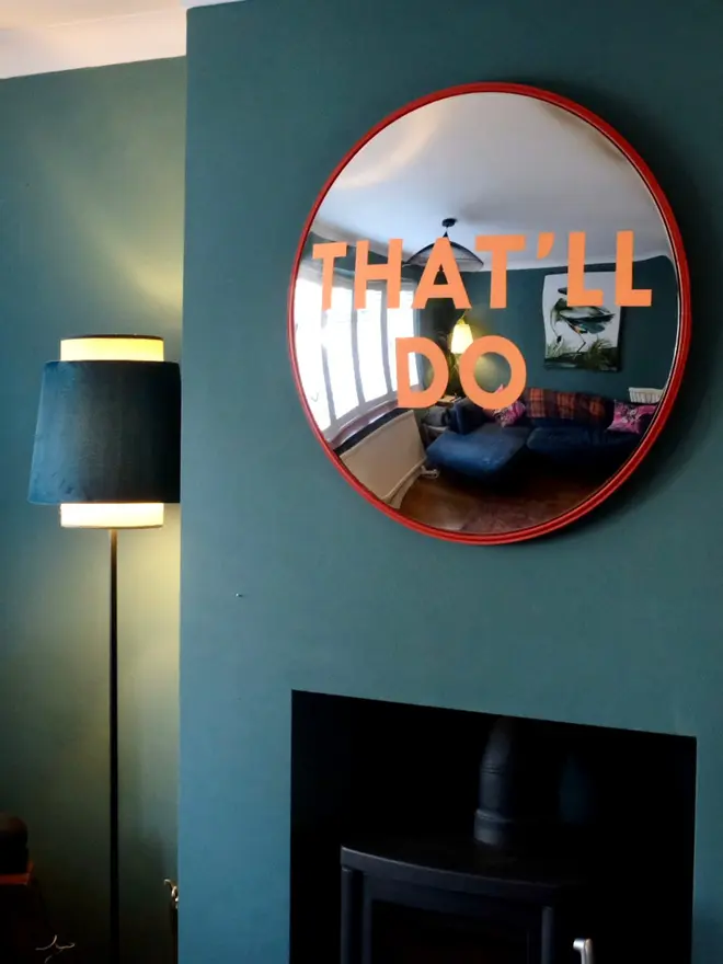 Round convex mirror with 'That'll Do' hand painted in orange in a red round frame, on a teal wall and log burner, with a lamp, logs and white ceiling