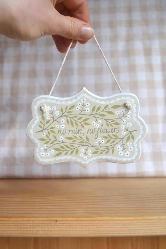 hanging clay sign with floral design reading no rain, no flowers' in front of gingham background