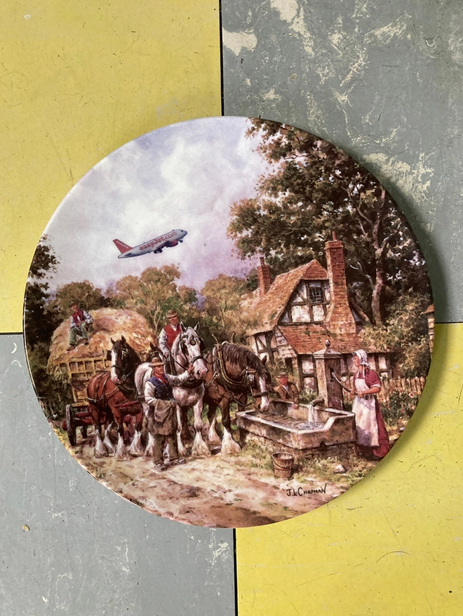 Decorative 8” plate featuring a chocolate box-twee cottage and a hay cart pulled by shirehorses. An easyJet plane soars off in the distance, causing a jarring contrast to the 17th century rural scene. 