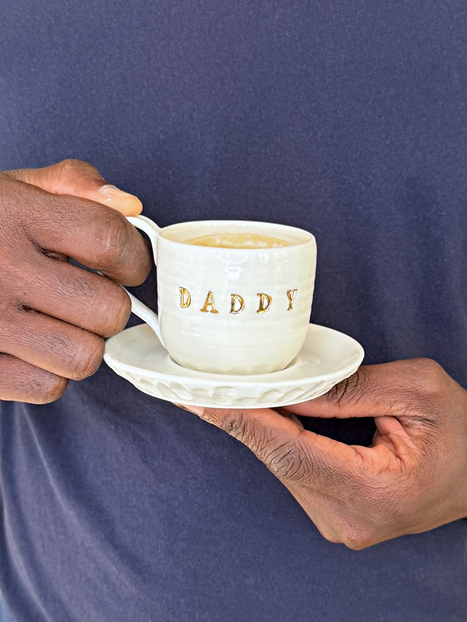 hand made porcelain espresso cup with gold lustre detailing on the text held in hands against navy blue background