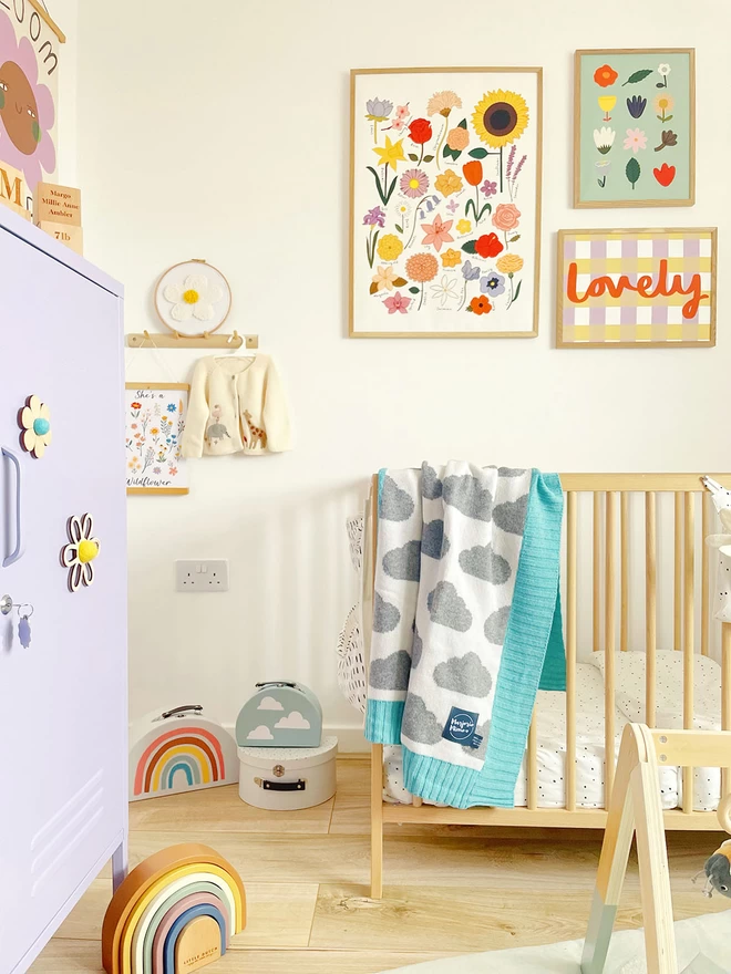 View of a bright and colourful baby's nursery. There is a baby gym on the floor in the foreground and a cot with the cloud blanket draped over the side in the background. Colourful prints of flowers hang behind the cot.