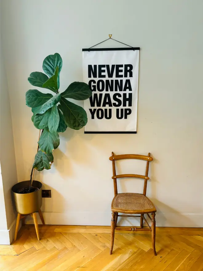 White tea towel with black text that says NEVER GONNA WASH YOU UP hung on wall with poster hanger frame and wooden/rattan chair and large plant in brass pot in front of it 