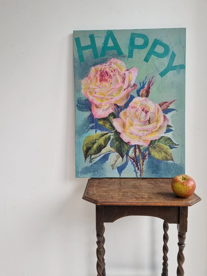 A painted plywood panel in turquoise and blue featuring decoupage of large pink roses taken from a Victorian seed packet with a motivational phrase that ‘happy’.