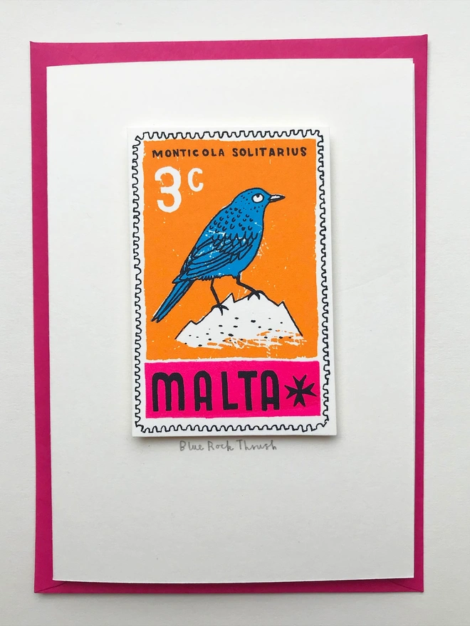 Bird Stamp Card Malta/ Blue Rock Thrush, a white card with a colourful bird design in the centre on top of a pink envelope on a white backdrop. 