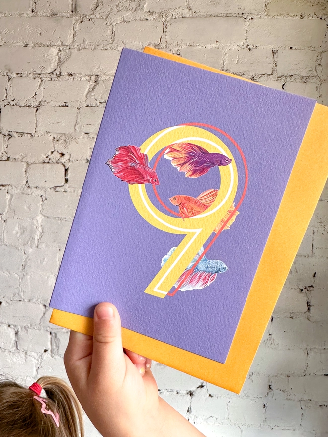 bright purple card with a big yellow number 9 and three bright illustrated betta fish with a bright yellow envelope