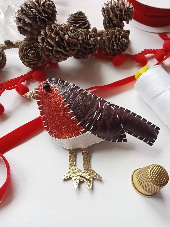 A leatherette robin brooch, hand stitched, with red glittery breast, sits amongst pine cones, red ribbon, pom pom trim, thimble and white reel of cotton