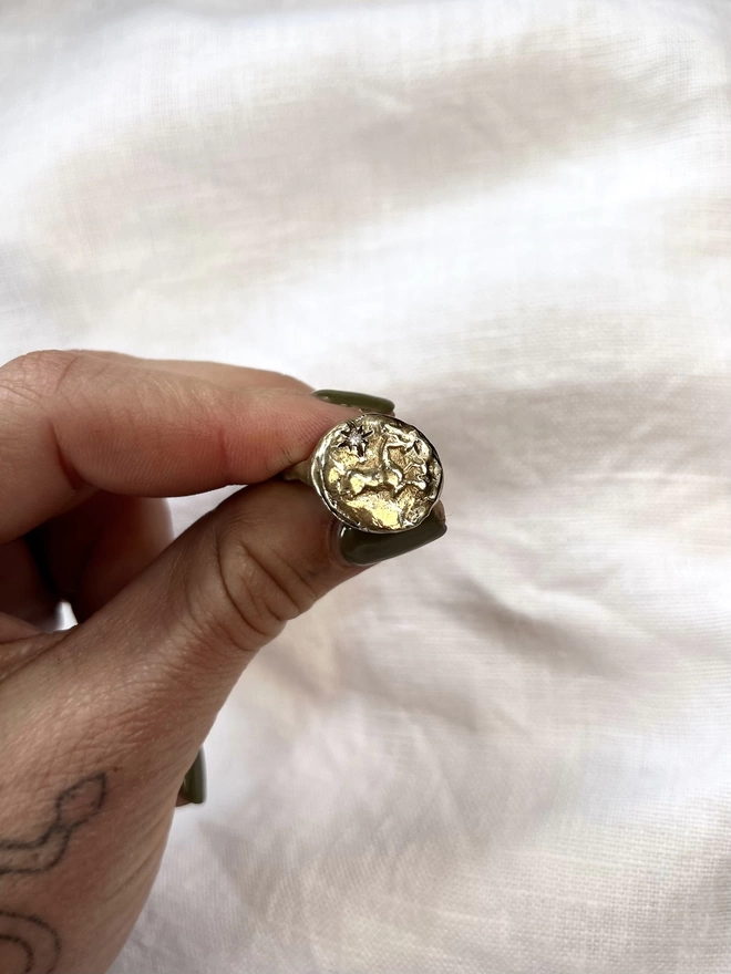 Image of a gold toned brass ring being held between the finer and thumb of a female hand, the ring has the image of a horse and star on it and the background is a piece of white linen fabric 
