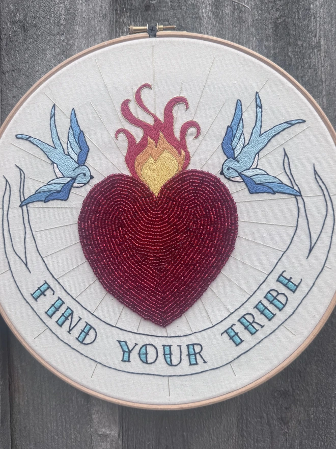 An embroidery hoop containing a red beaded heart with flames gild rays two bluebirds swallows and a banner containing the words find your tribe 