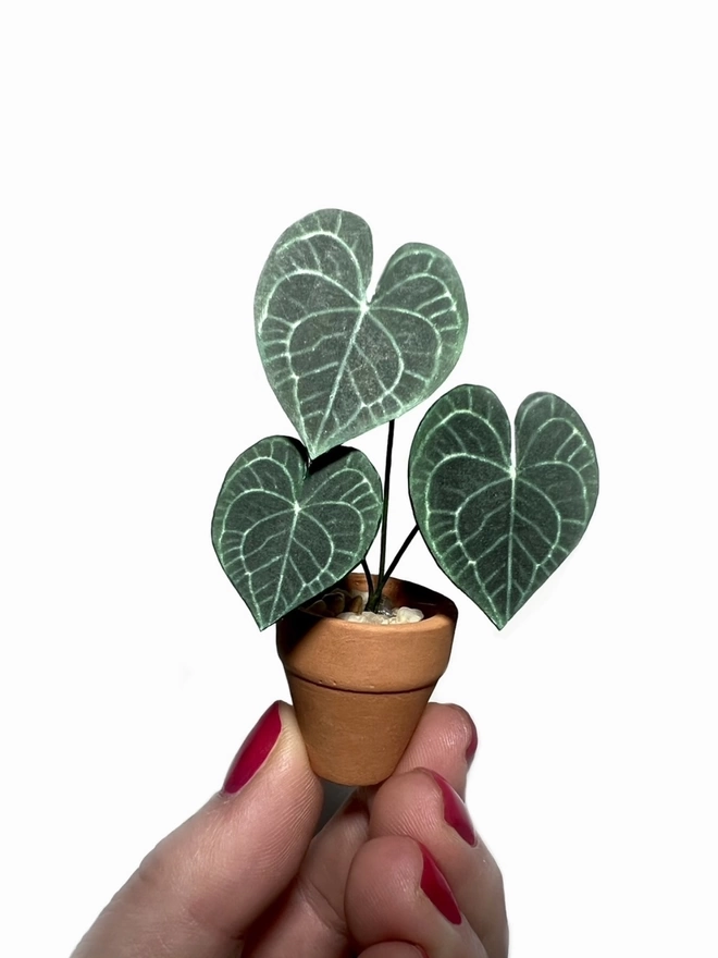 A miniature replica Anthurium Clarinervium paper plant ornament in a terracotta pot being held between 2 fingers against a white background