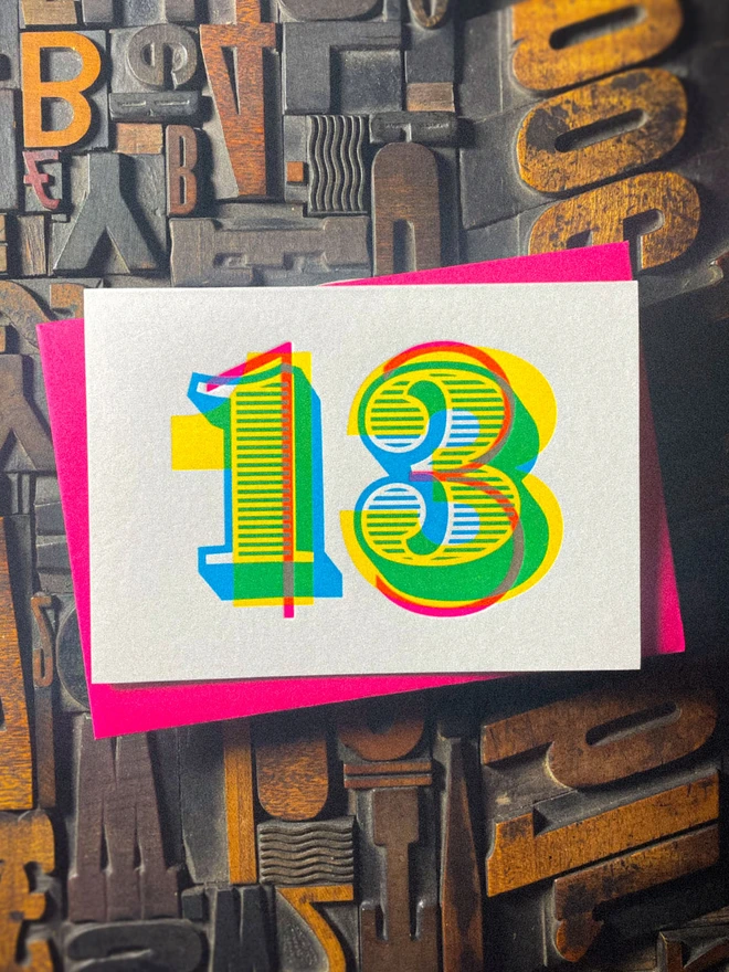 Colourful handmade 13th birthday letterpress card paired with a vibrant pink envelope. The oversized number 13 combines overlapping cyan, magenta and yellow inks, individually hand printed using traditional letterpress techniques.