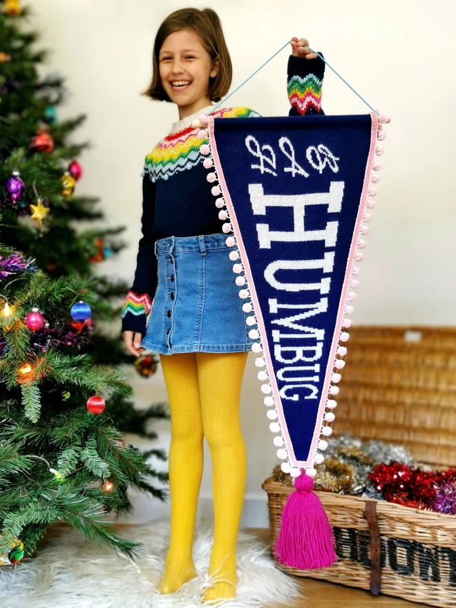 A girl wearing a colourful Christmas fairisle jumper stands next to a Christmas tree with retro coloured baubles. She is holding up a navy knitted pennant flag with the words ‘Bah Humbug’ written in a white sparkly retro text down it. The banner is trimmed with blush pink pom pom trim and has an oversize raspberry pink tassel hanging from the bottom of it. The banner is large, almost as tall as the girl! 