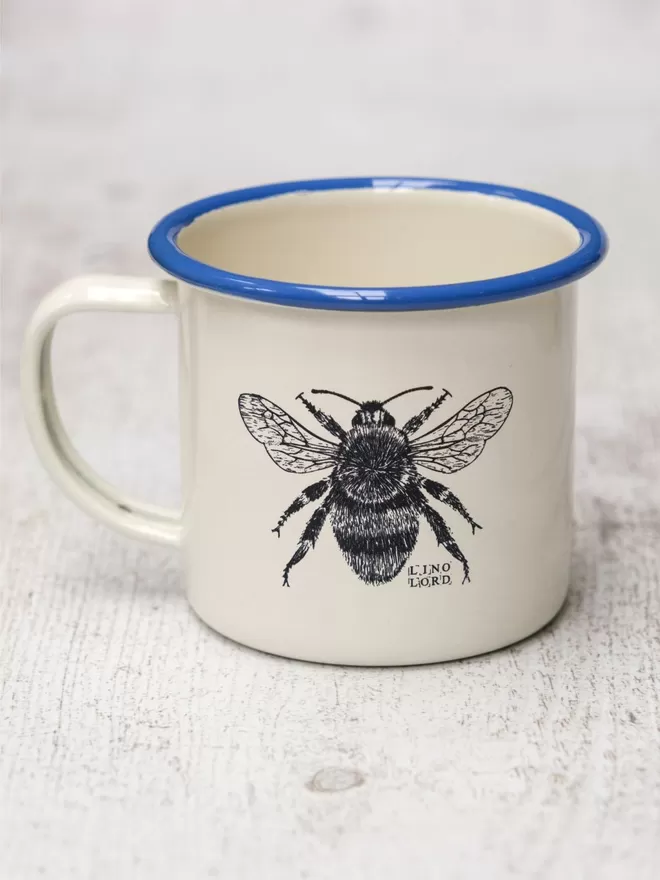 Picture of a Cream Enamel Mug with a Blue Rim with a Bee design etched onto it, taken from an original Lino Print