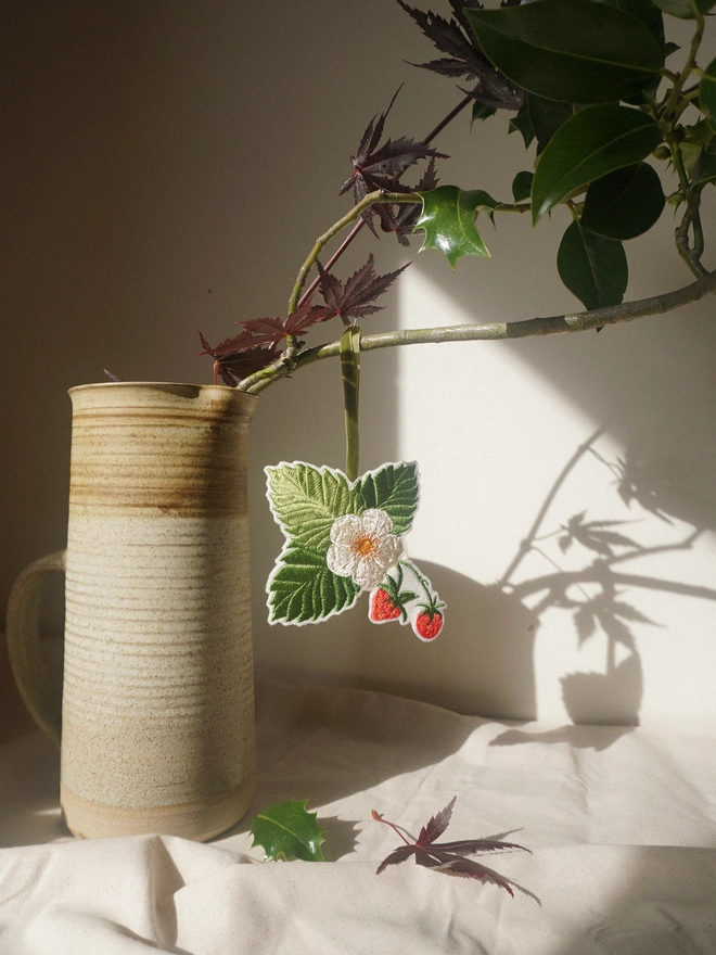 Embroidered strawberry and blossom hanging decoration with green velvet ribbon, styled with seasonal foliage.